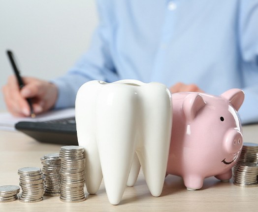 A ceramic model of a tooth, a piggy bank, and stacks of coins on a wooden table