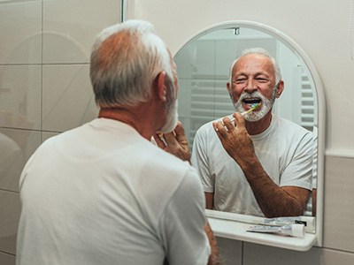 An older, cheerful man brushing his teeth in front of a bathroom mirror
