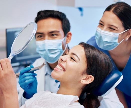 A woman admiring her smile with a hand mirror while dentists stand behind her